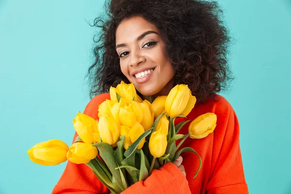 Gorgeous american woman with messy hair looking on camera and holding bouquet of beautiful yellow flowers isolated over blue background