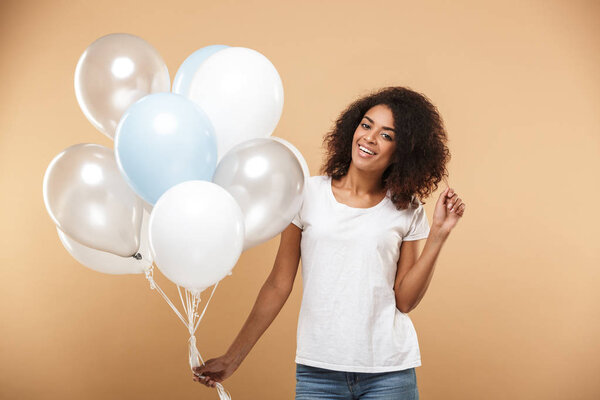 Portrait of a lovely young african woman holding bunch of air balloons isolated over beige background