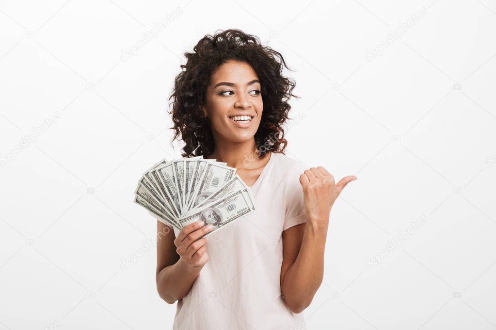 Lucky american woman with curly brown hair holding fan of money dollar banknotes and gesturing finger aside on copyspace isolated over white background