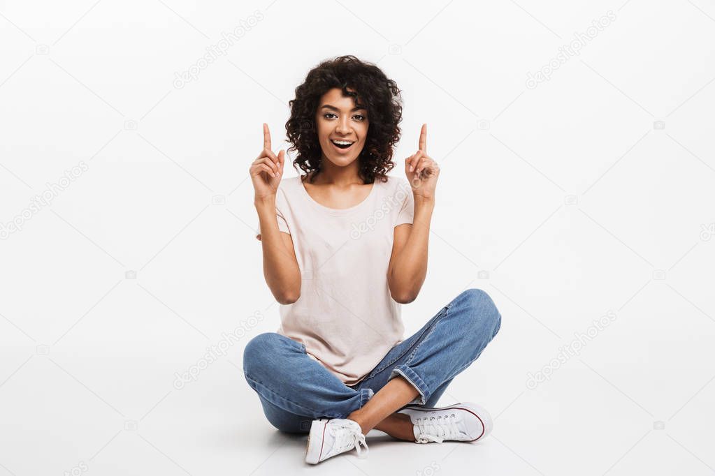 Portrait of cheerful young afro american woman pointing fingers up while sitting on a floor with legs crossed isolated over white background