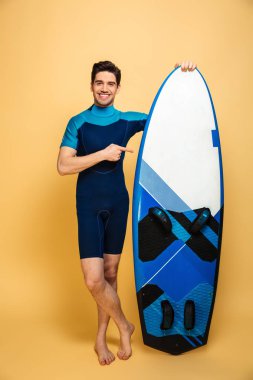 Yakışıklı bir genç adam portresi görünümlü kamera işaret sarı arka plan üzerinde izole surfboard holding mayo giymiş.