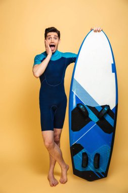 Heyecanlı bir genç adam portresi tam uzunlukta sarı arka plan üzerinde izole surfboard holding mayo giymiş