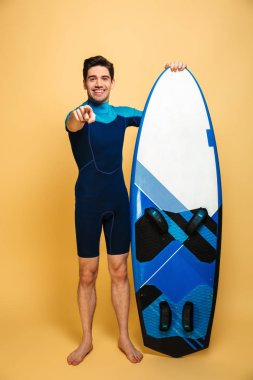 Heyecanlı bir genç adam portresi tam uzunlukta surfboard holding ve sarı arka plan üzerinde izole kamera, parmak işaret mayo giymiş