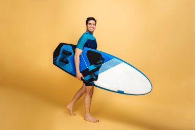 Mutlu bir genç adam portresi sarı arka plan üzerinde izole surfboard holding mayo giymiş.