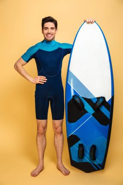 Genç gülümseyen bir adam portresi tam uzunlukta sarı arka plan üzerinde izole surfboard holding mayo giymiş