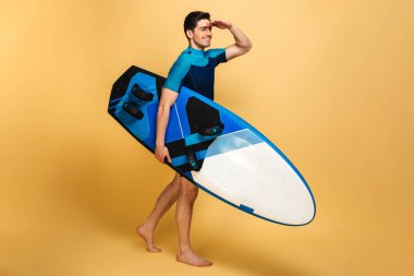 Heyecanlı bir delikanlı imajını sarı arka plan bir kenara inceliyorlar izole surfboard holding mayo giymiş.