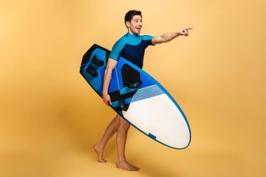 Heyecanlı bir delikanlı imajını bir yana işaret arıyorum sarı arka plan üzerinde izole surfboard holding mayo giymiş.