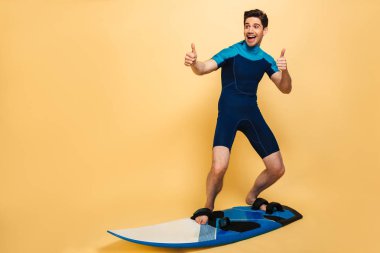 Mutlu bir genç adam portresi tam uzunlukta bir gemide sörf ve başparmak sarı arka plan üzerinde izole gösterilmesini mayo giymiş