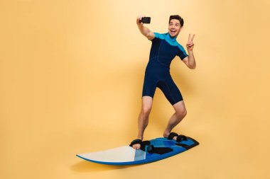 Mutlu bir genç adam portresi tam uzunlukta sarı arka plan üzerinde sörf tahtası izole iken bir selfie alarak mayo giymiş