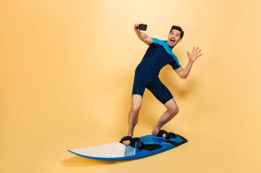 Sörf tahtası üzerinde sarı arka plan izole iken bir selfie alarak mayo giymiş bir heyecanlı genç adam tam uzunlukta portresi