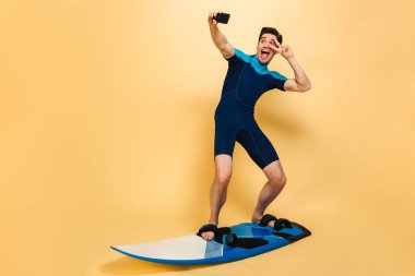 Genç gülümseyen bir adam portresi tam uzunlukta sarı arka plan üzerinde sörf tahtası izole iken bir selfie alarak mayo giymiş