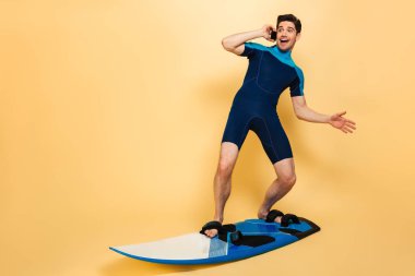 Kendine güvenen genç bir adam portresi tam uzunlukta sarı arka plan üzerinde sörf tahtası izole süre üstünde hareket eden telefon konuşuyor mayo giymiş