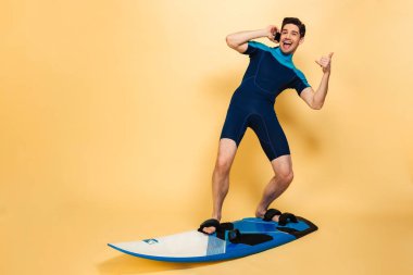 Fotoğraf heyecanlı genç bir adam üzerinde sörf tahtada telefonla konuşurken sarı arka plan izole mayo giymiş.