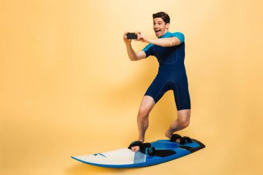 Kendine güvenen genç bir adam portresi tam uzunlukta sarı arka plan üzerinde sörf tahtası izole süre cep telefonunuzla resim konuşuyor mayo giymiş