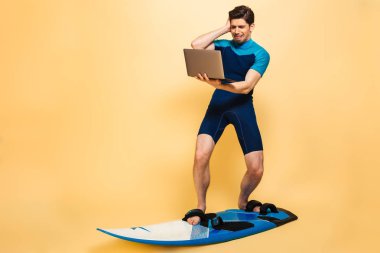 Üzgün bir genç adam portresi tam uzunlukta sarı arka plan üzerinde sörf tahtası izole dizüstü bilgisayar tutarken mayo giymiş