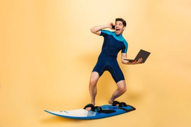 Dizüstü bilgisayar kullanan telefonla konuşurken sörf tahtası üzerinde sarı arka plan üzerinde izole mayo fotoğraf heyecanlı bir gencin giymiş.