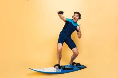 Fotoğraf sörf tahtası üzerinde sarı arka plan üzerinde izole mayo giymiş bir duygusal delikanlı olun selfie tarafından telefon kulaklık ile müzik dinleme.