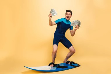 Mutlu bir genç adam portresi tam uzunlukta sarı arka plan üzerinde sörf tahtası izole para banknot demet tutarken mayo giymiş