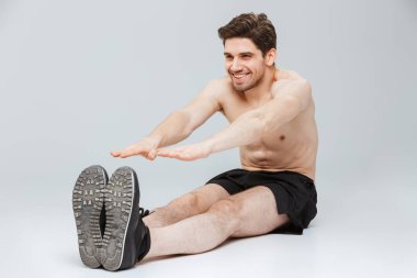 Gri arka plan üzerinde izole otururken germe egzersizleri yapıyor bir gülümseyen genç yarı çıplak sporcu portresi