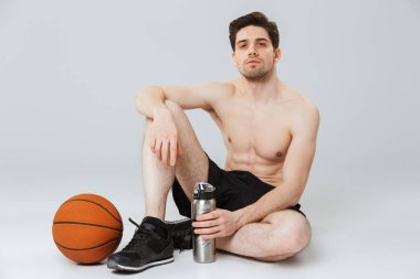 Gri arka plan üzerinde izole basketbol ile otururken su şişesi tutan bir kendine güvenen genç yarı çıplak sporcu portresi