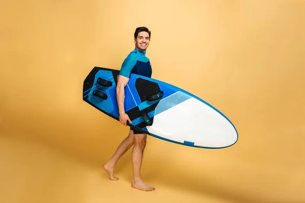 黄色の背景に分離したサーフボードを持って水着に身を包んだ幸せな若い男の肖像 — ストック写真