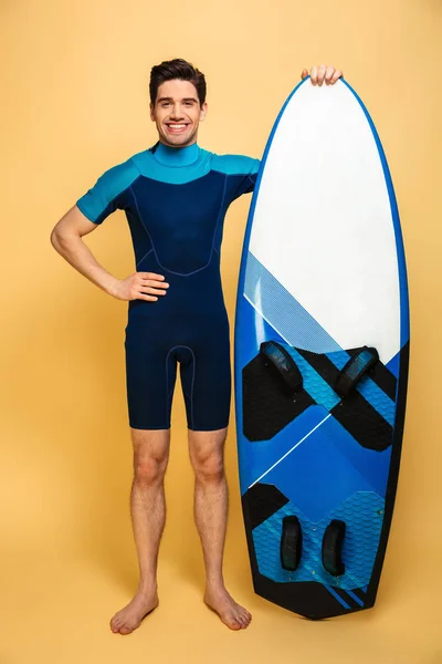 黄色の背景に分離したサーフボードを持って水着に身を包んだ笑顔若い男の完全な長さの肖像画 — ストック写真