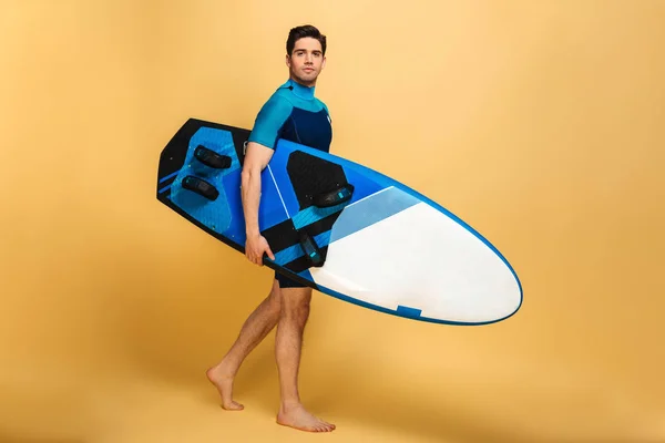 黄色の背景に分離したサーフボードを持って水着に身を包んだハンサムな若い男の肖像 — ストック写真
