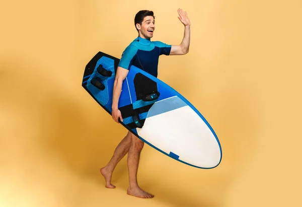 脇を振って見て黄色背景に分離したサーフボードを持って水着に身を包んだ興奮して若い男のイメージ — ストック写真