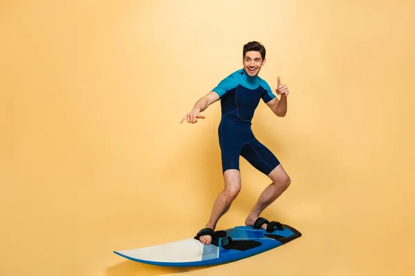 黄色背景に分離された基板のサーフィン水着に身を包んだ陽気な若い男の完全な長さの肖像画 — ストック写真