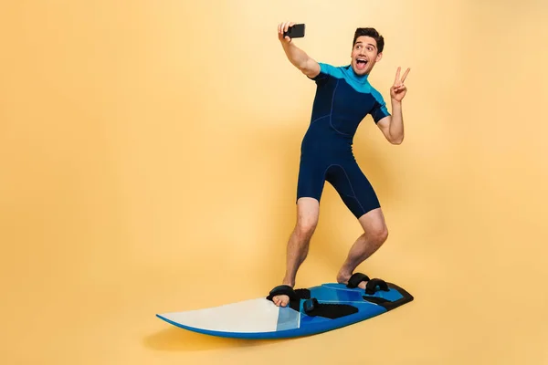 黄色の背景に分離されたボードでサーフィンしながら Selfie を取って水着に身を包んだ陽気な若い男の完全な長さの肖像画 — ストック写真
