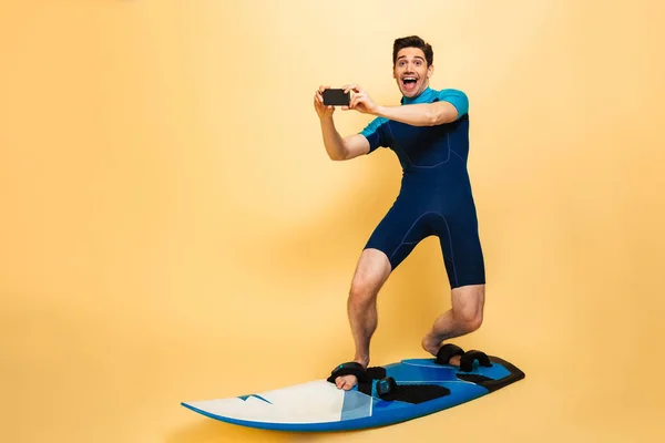 黄色の背景に分離されたボードでサーフィンしながら携帯電話で写真を撮って水着に身を包んだ興奮して若い男の完全な長さの肖像画 — ストック写真