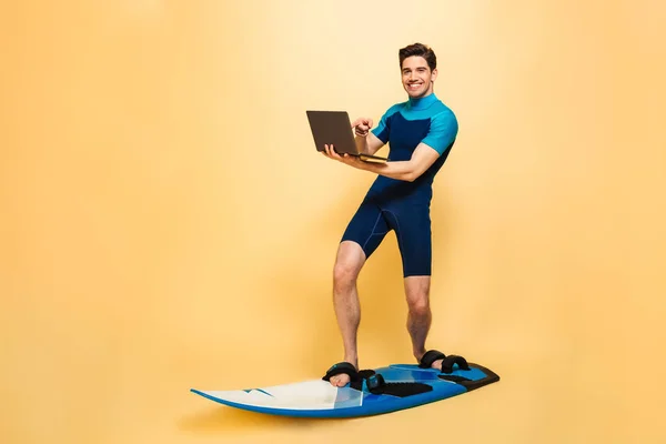 幸せな若い男の完全な長さの肖像画は黄色背景に分離されたボードでサーフィン中にラップトップ コンピューターを指して水着に身を包んだ — ストック写真