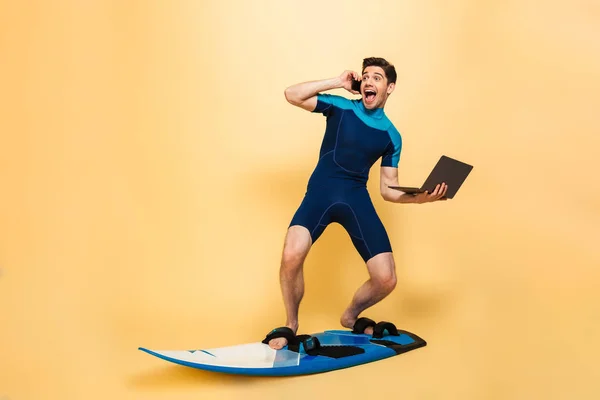 ラップトップ コンピューターを使用して電話で話しサーフィン ボードの背景が黄色で分離された水着を着て興奮して若い男の写真 — ストック写真