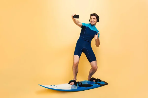 サーフィン ボードの黄色背景に分離した水着に身を包んだ感情的な若い男の写真を行う携帯電話のヘッドフォンで音楽を聴く Selfie — ストック写真