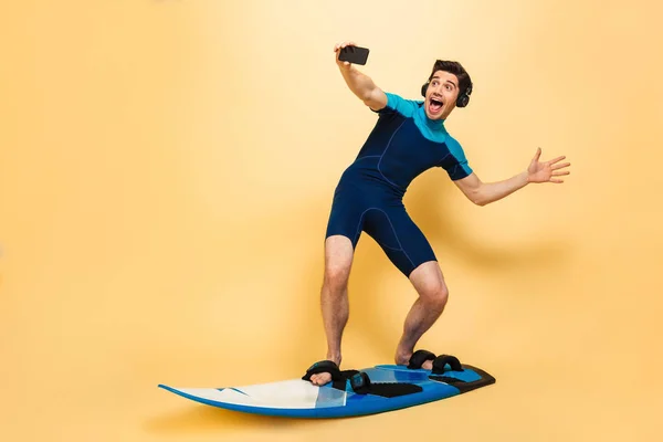 サーフィン ボードの黄色背景に分離した水着に身を包んだ若い男の写真を行う携帯電話のヘッドフォンで音楽を聴く Selfie — ストック写真