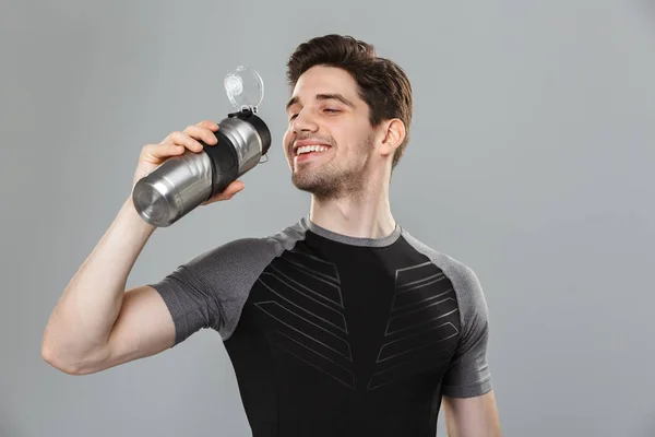 一个微笑的年轻运动员的肖像在灰色背景下被隔离的水瓶饮用 — 图库照片