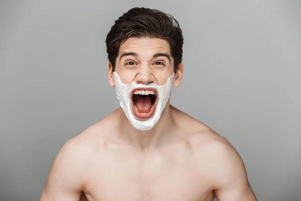 Beauty portrait of half naked handsome young man with shaving foam on his face screaming isolated over gray background