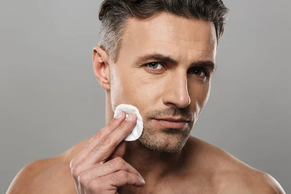 Image of serious concentrated mature man standing isolated over grey wall background naked take care of his skin. Looking camera.