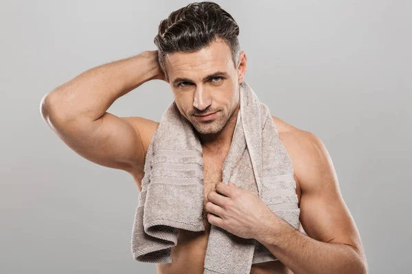 Picture of handsome mature man standing isolated over grey wall background naked. Looking camera holding towel.