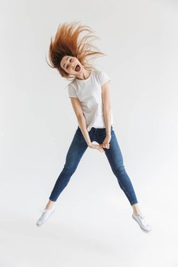 Tam boy görüntü eğleniyor atlama ve kamera seyir gri arka plan üzerinde tişört içinde neşeli çığlık atan kadın