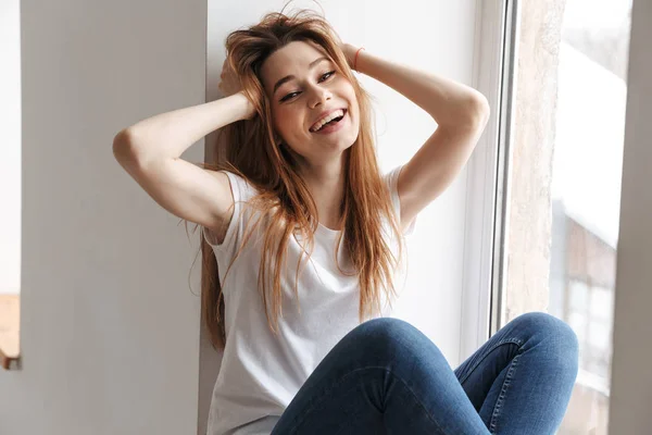 Mujer Alegre Camiseta Sentada Alféizar Ventana Mientras Sostiene Cabeza Mira — Foto de Stock