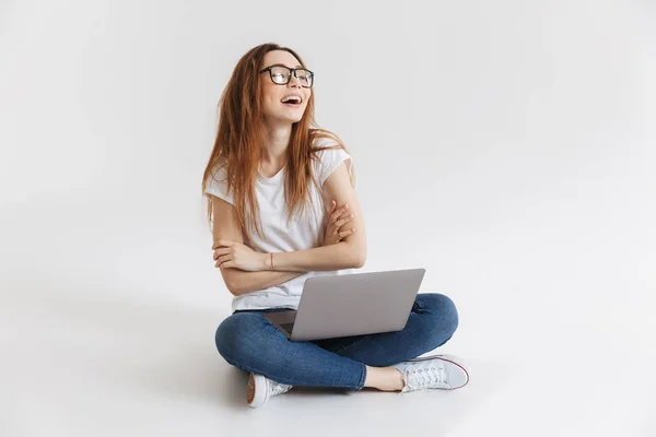 T恤和眼镜的开朗女人坐在地板上用笔记本电脑 同时手持交叉双臂 仰望灰色背景 — 图库照片
