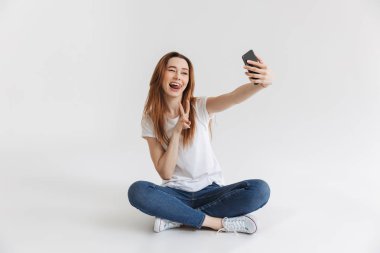 Bacaklar ile otururken selfie cep telefonu ile alarak mutlu bir genç kız portresi izole beyaz arka plan üzerinde çarpı işareti