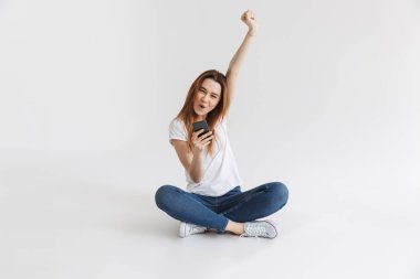 Bacaklar ile otururken cep telefonuyla neşeli bir genç kız portresi izole beyaz arka plan üzerinde çarpı işareti