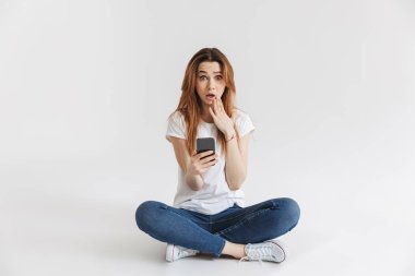 Bacaklar ile otururken cep telefonuyla şok bir genç kız portresi izole beyaz arka plan üzerinde çarpı işareti