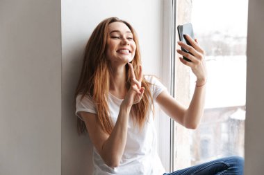 Pencere kenarında oturan ve selfie barış hareketi gösterilen süre Smartphone'da yapım t-shirt mutlu kadın