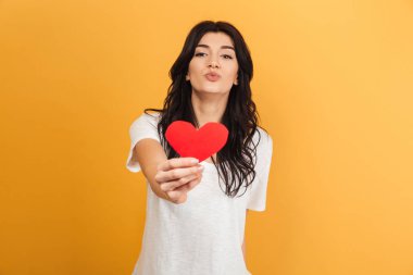 Görüntü izole kalp tutan sarı arka plan üzerinde duran genç Sevimli güzel kadın.