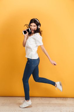 Görüntü izole cep telefonu tutan müzik şarkı dinleme sarı arka plan üzerinde dans atlama genç güzel kadın.