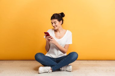 Bacaklar ile otururken cep telefonuyla genç gülümseyen bir kadın portresi bir yere sarı arka plan üzerinde geçti.