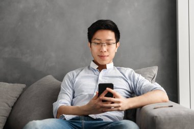 Evde bir kanepede otururken cep telefonuyla gülümseyen genç bir Asyalı adam portresi